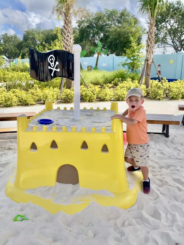 A young boy standing next to a yellow sandcastle structure with a pirate flag in a sunny outdoor setting. Pirate Island Sand Play