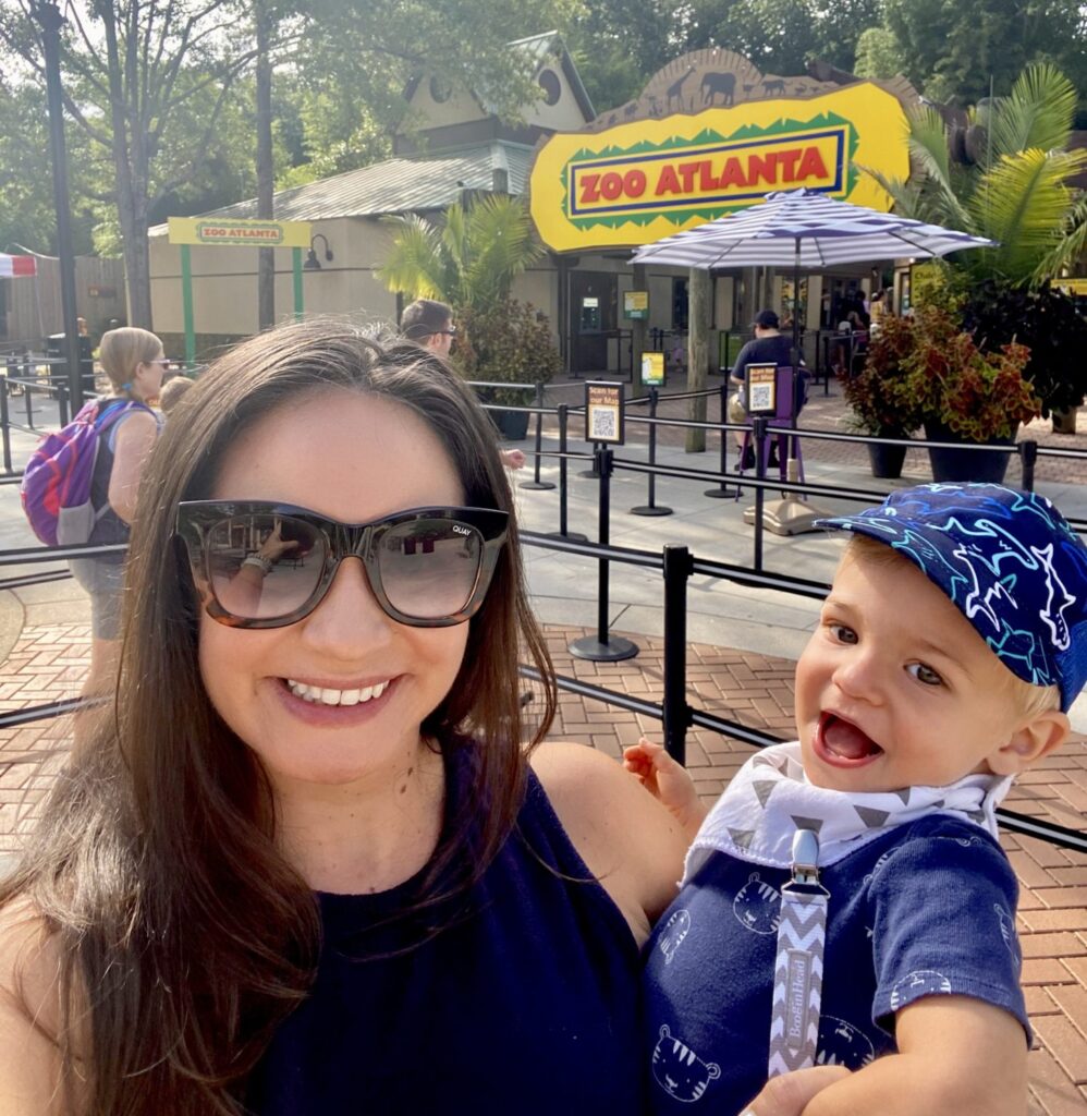 A woman holding a toddler outside the Zoo Atlanta entrance, both smiling with people and signage in the background.