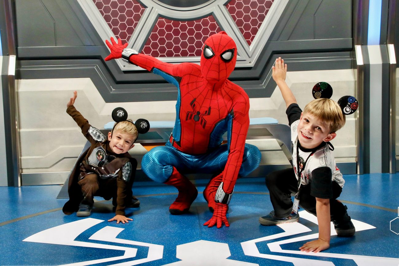 Two children in themed costumes posing with Spider-Man in a superhero-themed setting, smiling and striking playful poses.