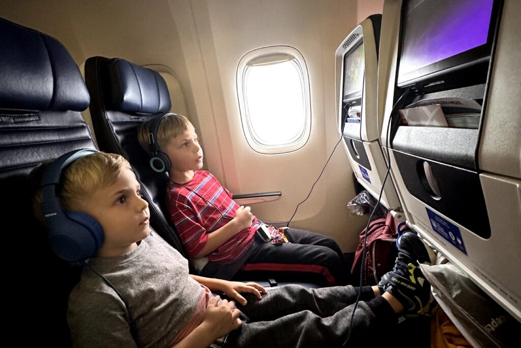 Two young children watching in-flight entertainment screens on an airplane, wearing headphones and looking engaged.