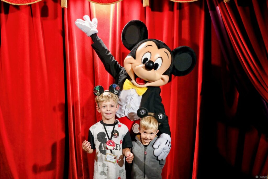 Two children posing with Mickey Mouse at Disney World, with red curtains in the background and wearing themed Mickey Mouse hats.