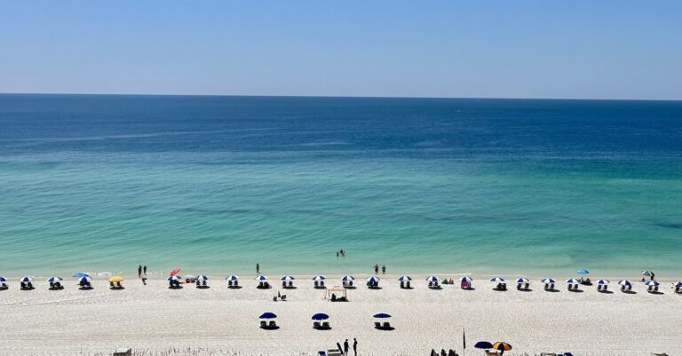 Aerial view of Pensacola Beach with rows of blue and white umbrellas lining the soft, white sand and people enjoying the clear, calm turquoise waters of the Gulf of Mexico.