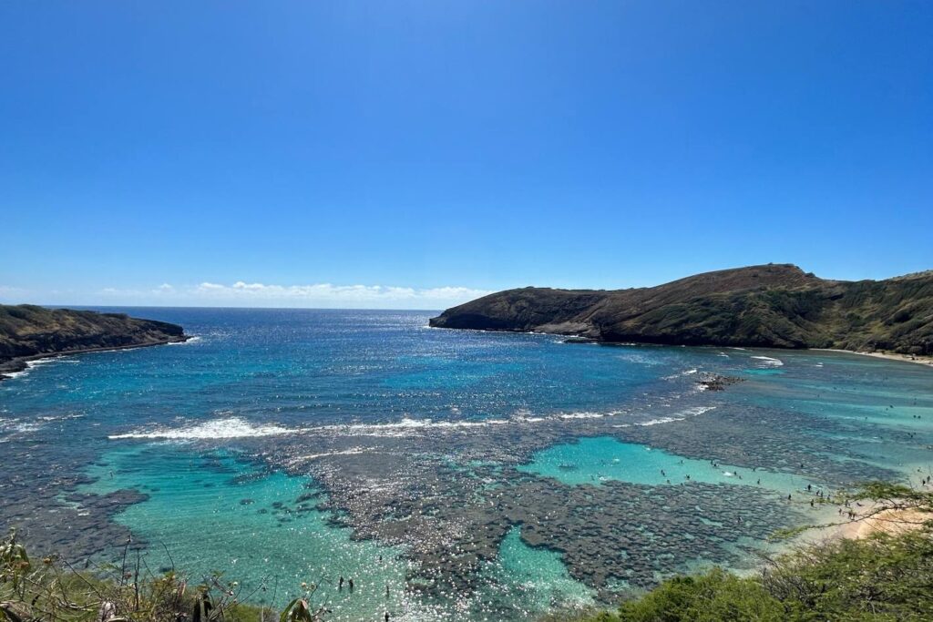 A panoramic view of Hanauma Bay in Oahu, Hawaii, featuring blue waters, visible coral reefs, and swimmers, surrounded by hills under a clear blue sky.