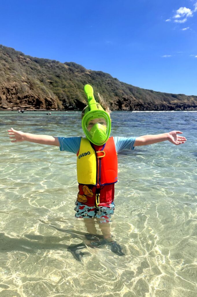 A young child wearing a green snorkel mask, a yellow and red life vest, and colorful swim shorts stands in the clear, shallow waters of Hanauma Bay in Oahu, Hawaii with arms outstretched, under a bright blue sky.