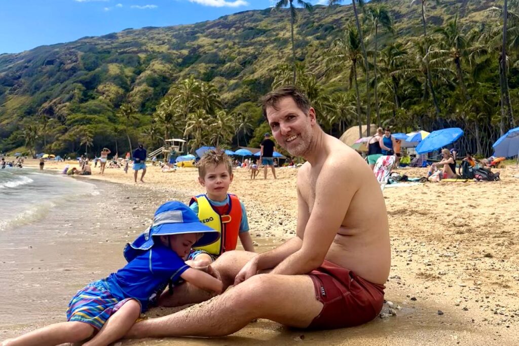 A father and two young children sit on the sandy beach at Hanauma Bay in Oahu, Hawaii, with green hills and palm trees in the background under a clear blue sky.