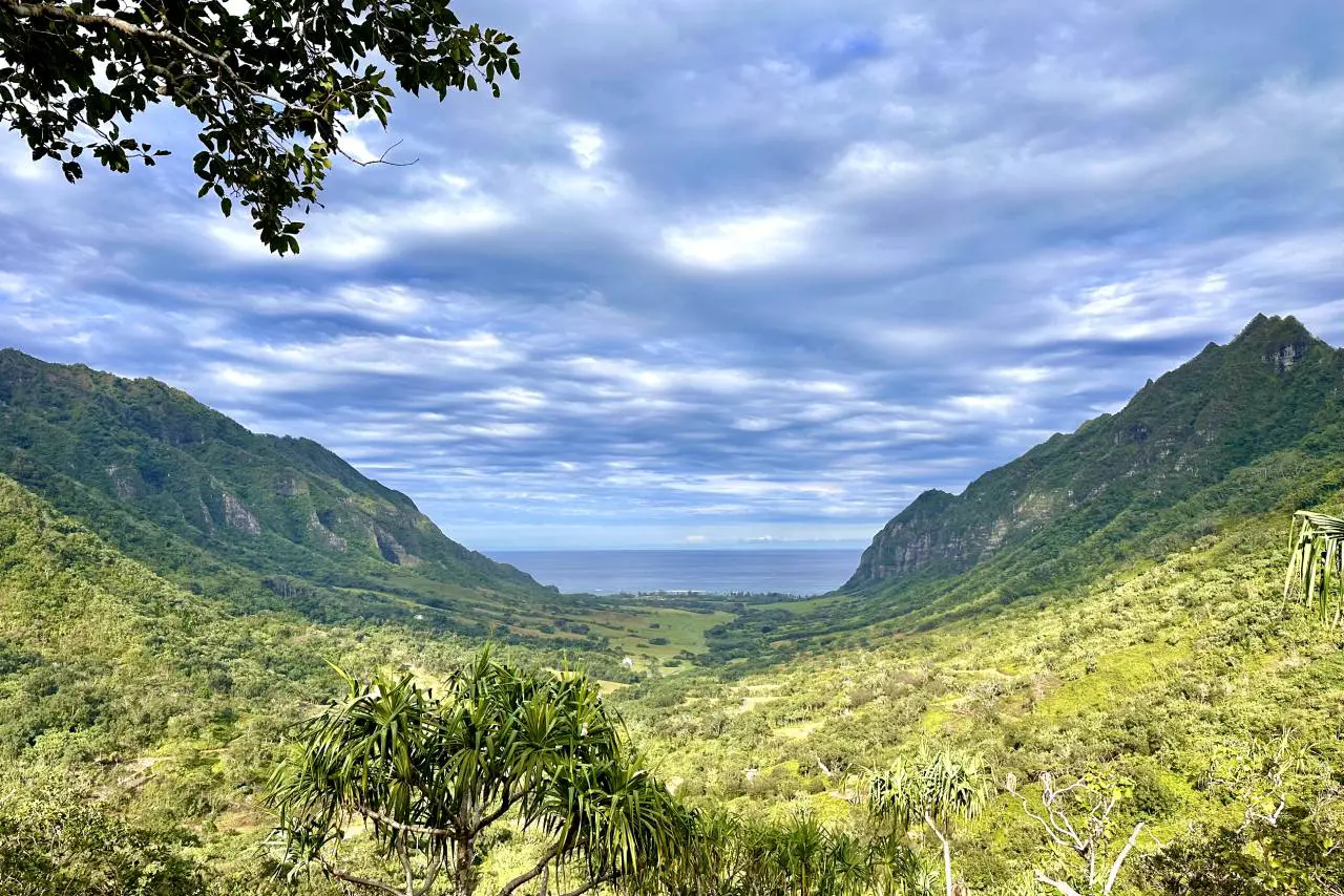 A panoramic view of a green valley surrounded by steep, forested mountains, with the blue ocean visible in the distance under a cloudy sky at Kualoa Ranch in Oahu.