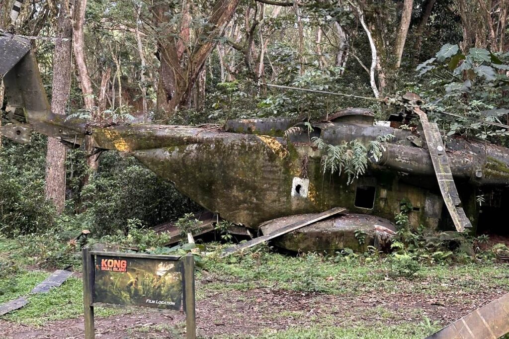 A close-up of a moss-covered, rusted helicopter prop from the movie "Kong: Skull Island" resting in a forested area, with a sign in the foreground that reads "Kong Skull Island Film Location," at Kualoa Ranch in Oahu.