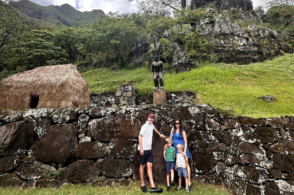 A family of four poses in front of a stone wall with a thatched-roof hut and a wooden statue behind them at Kualoa Ranch in Oahu. The parents stand with their two young children, all smiling for the photo.