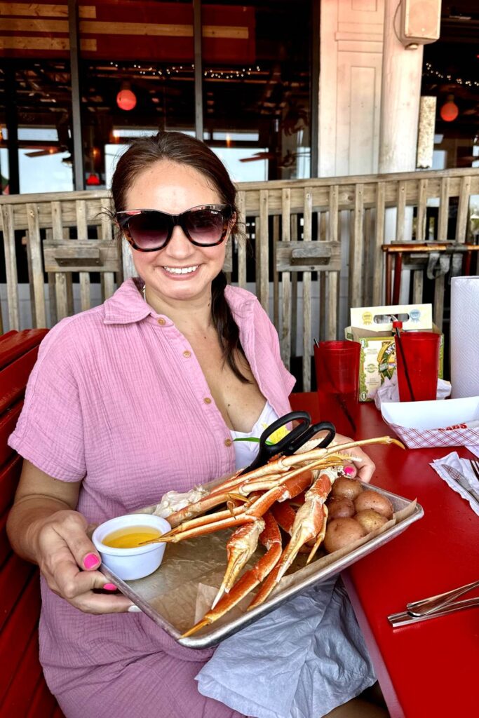 A woman in a pink shirt and sunglasses smiles while holding a tray of crab legs in, boiled potatoes, and a small cup of melted butter at Crabs Pensacola Beach restaurant.