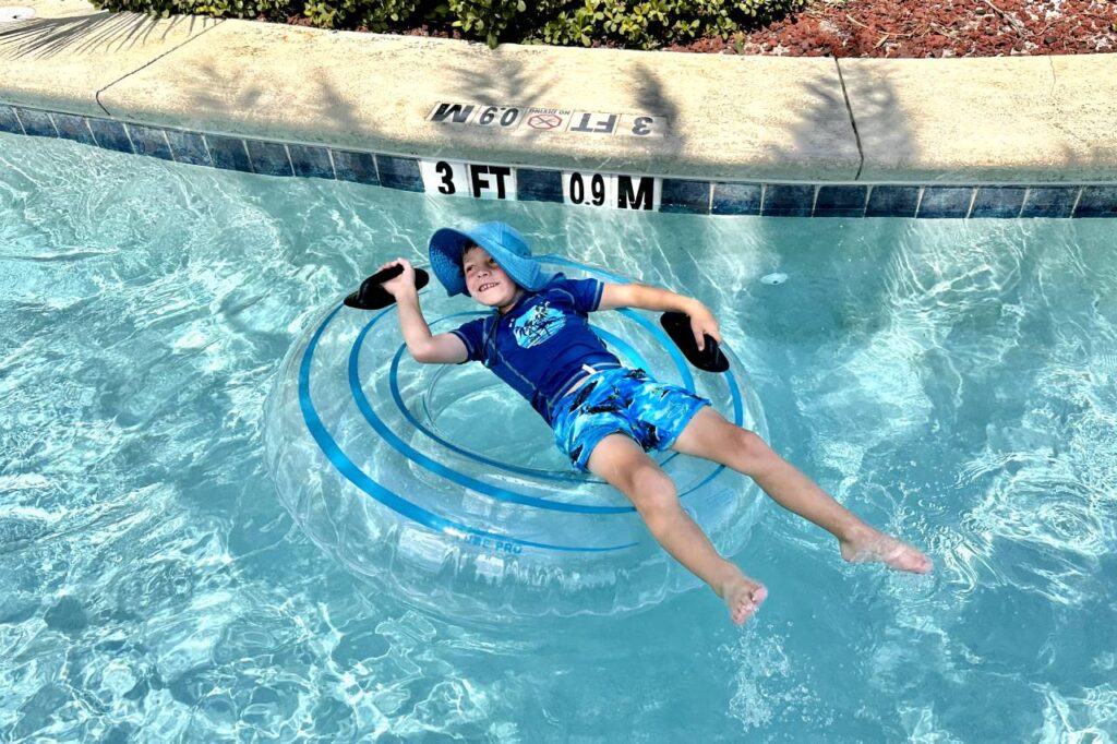 A young boy wearing a blue sun hat and swimwear floats happily in a clear inflatable tube in a swimming pool in The Holiday Inn Resort Pensacola Beach, with his feet in the water and a big smile on his face. The pool is shallow, as indicated by the "3 FT" depth marker on the edge.