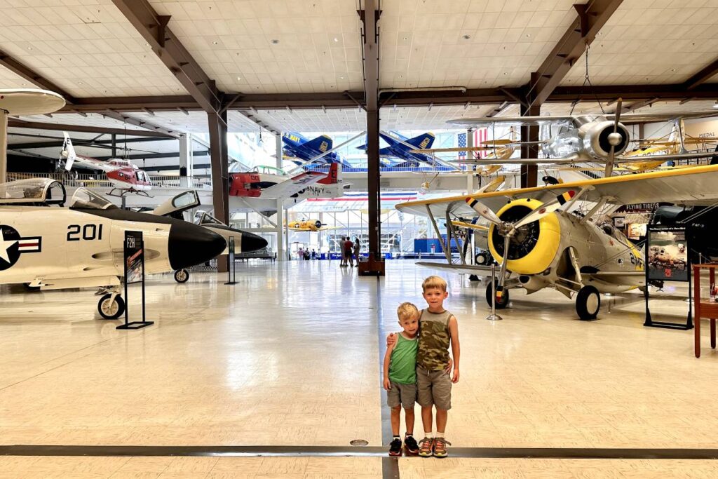 Two young boys stand together, smiling in front of a display of vintage aircraft inside the National Naval Aviation Museum in Pensacola. The museum hangar is filled with various planes, including a yellow and silver biplane and a white jet with "201" on its side.