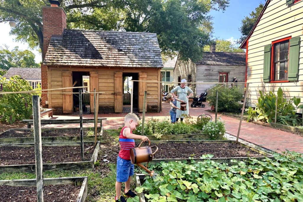 A young boy waters plants in a garden bed with a watering can, while another child, with the guidance of an adult dressed in period clothing, waters nearby. The garden is part of a historical village setting in Pensacola, with wooden buildings and a brick path visible in the background.