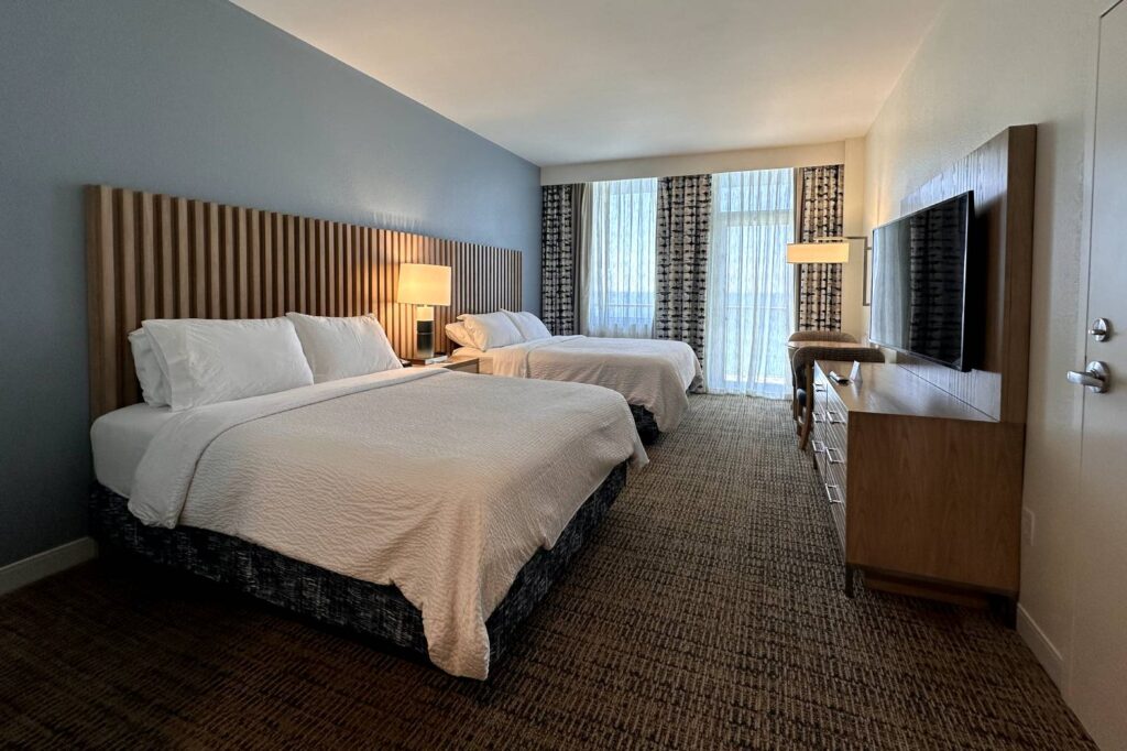 A Holiday Inn Resort Pensacola Beach room with two double beds, a large flat-screen TV on a wooden dresser, and a tall floor lamp near curtains that allow natural light to fill the room.