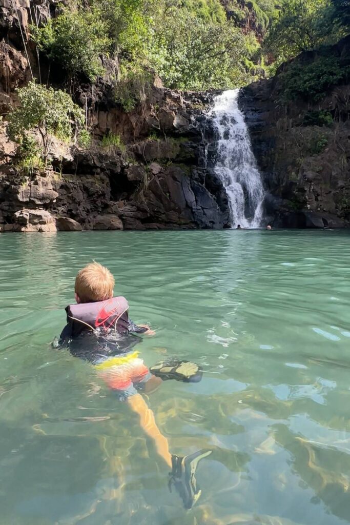 A young boy wearing a life jacket floats in a clear green pool while looking up at Waimea Falls in Oahu, Hawaii.