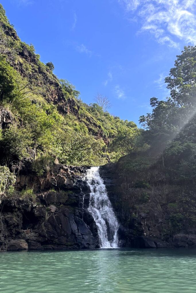 The beautiful Waimea Fall in Oahu cascades down a rocky cliff into a clear, green pool below, under a bright blue sky.