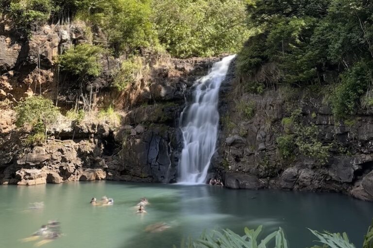 The beautiful Waimea Fall in Oahu, Hawaii cascades down a rocky cliff into a clear, green pool below.