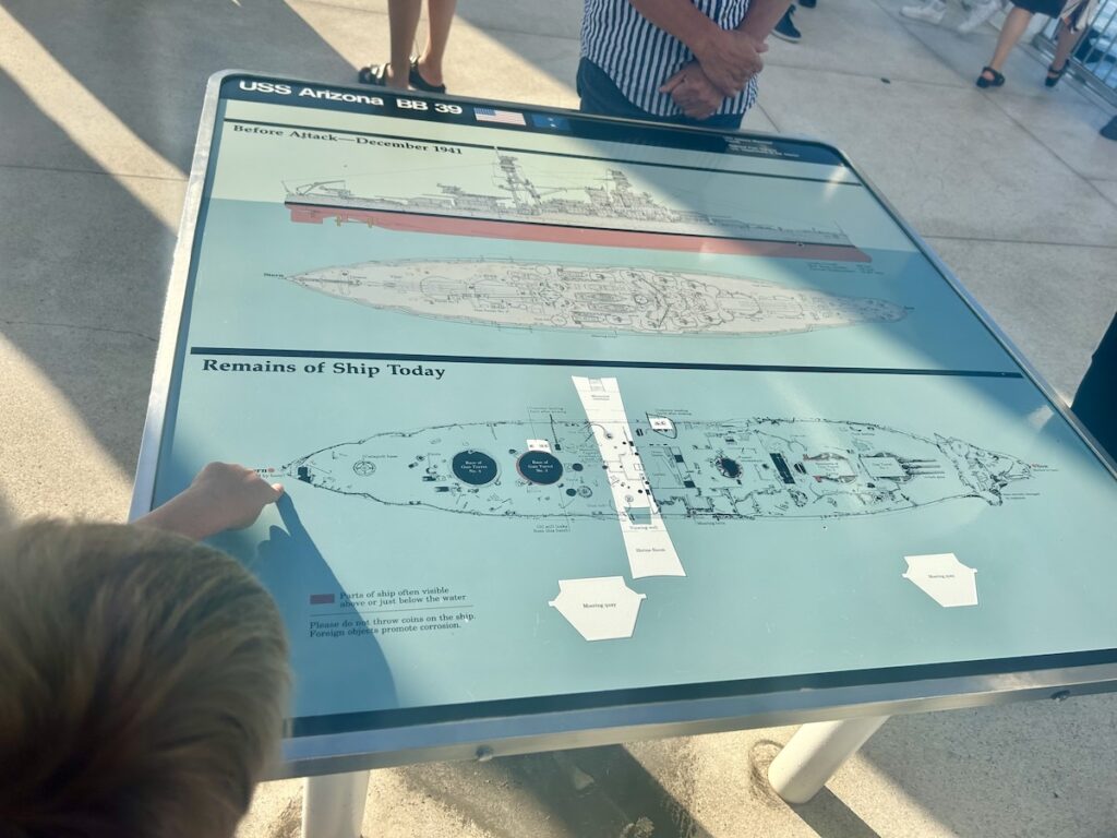 A child points to a display showing diagrams of the USS Arizona before its sinking and its current underwater remains. The sign offers a visual comparison, helping visitors understand the ship's structure. Pearl Harbor, Hawaii