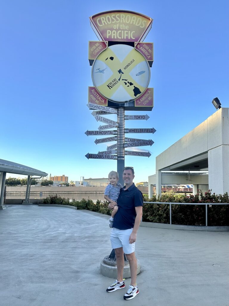 A man is holding a young child in his arms, both smiling, as they stand in front of a colorful signpost labeled "Crossroads of the Pacific." The sign features directional arrows pointing to various locations around the world from Pearl Harbor, Oahu, Hawaii.