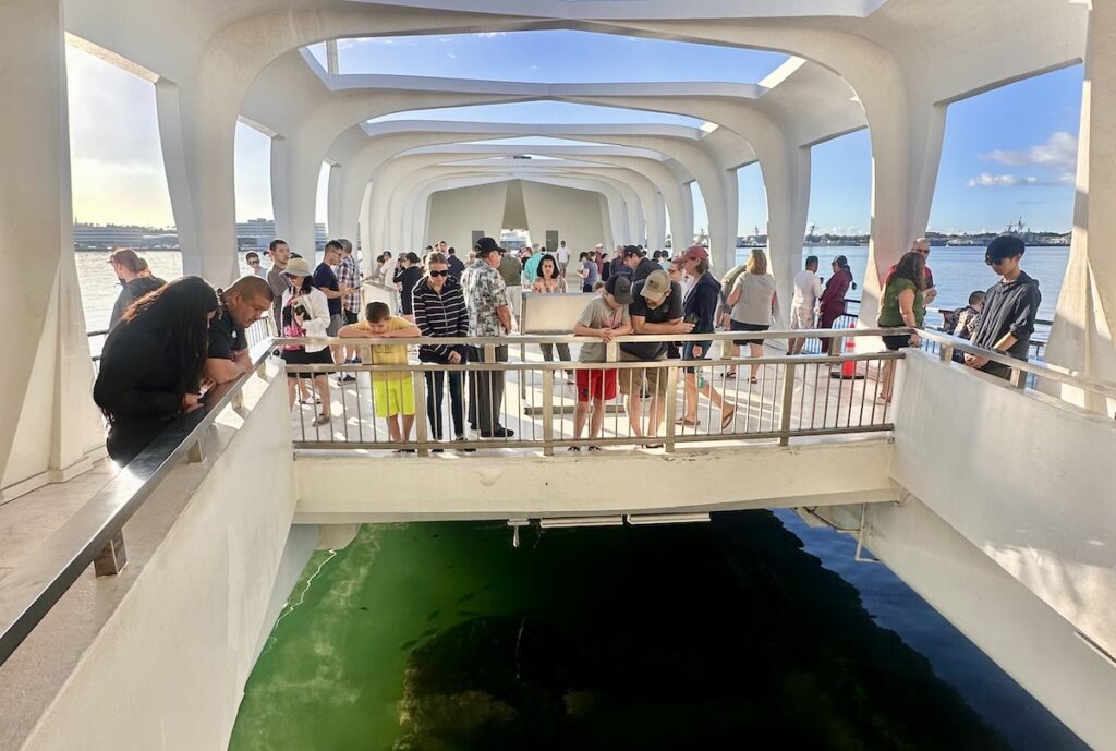 Visitors inside the USS Arizona Memorial lean over railings, looking down at the remains of the sunken battleship below. The white structure features open windows, allowing sunlight and views of the surrounding harbor. Pearl Harbor, Hawaii