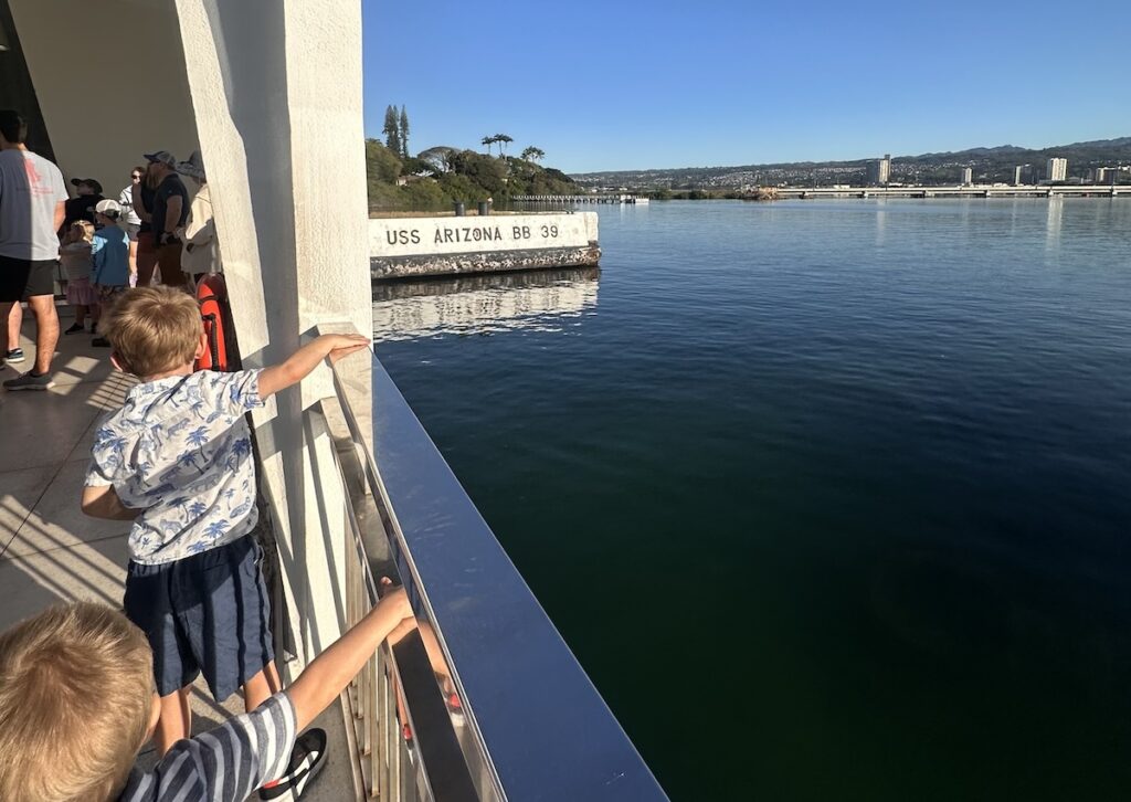 Two young boys look out over the railing at the USS Arizona Memorial, with one of them pointing toward the white structure marked "USS Arizona BB 39" floating in the water. Pearl Harbor, Oahu