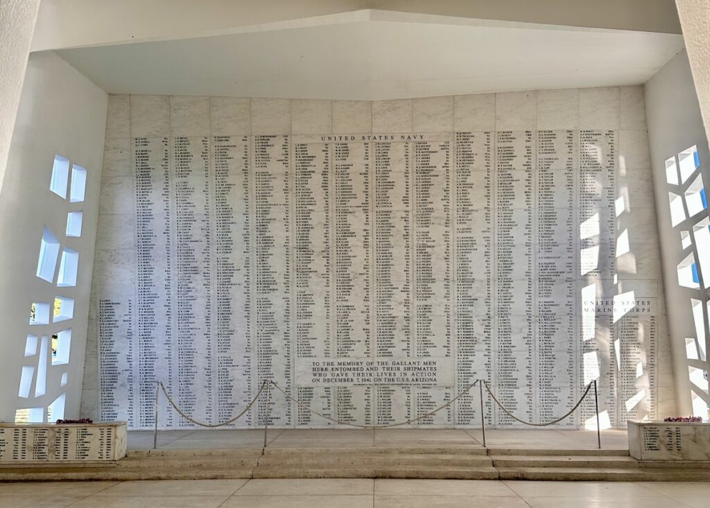 The image shows the interior of the USS Arizona Memorial, featuring a large white marble wall inscribed with the names of the 1,177 sailors and Marines who lost their lives during the attack on Pearl Harbor in Oahu.