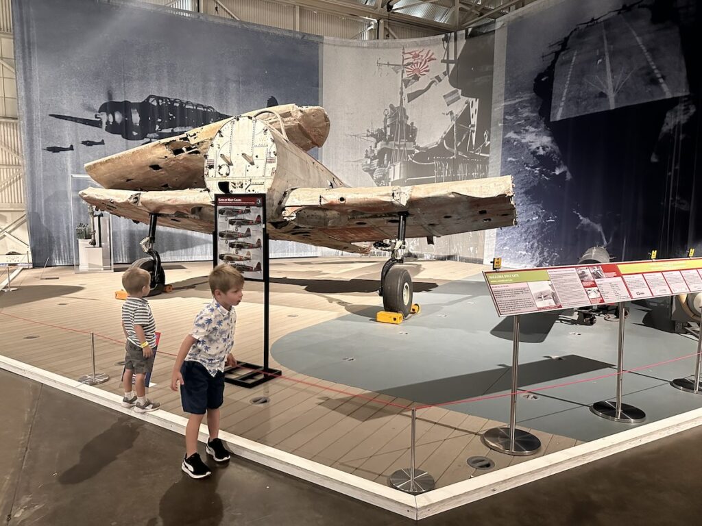 A young child observes a display featuring a damaged, historical aircraft inside the Pacific Aviation Museum. The plane is partially restored and surrounded by informational signs, offering insight into its past. Pearl Harbor, Oahu, Hawaii