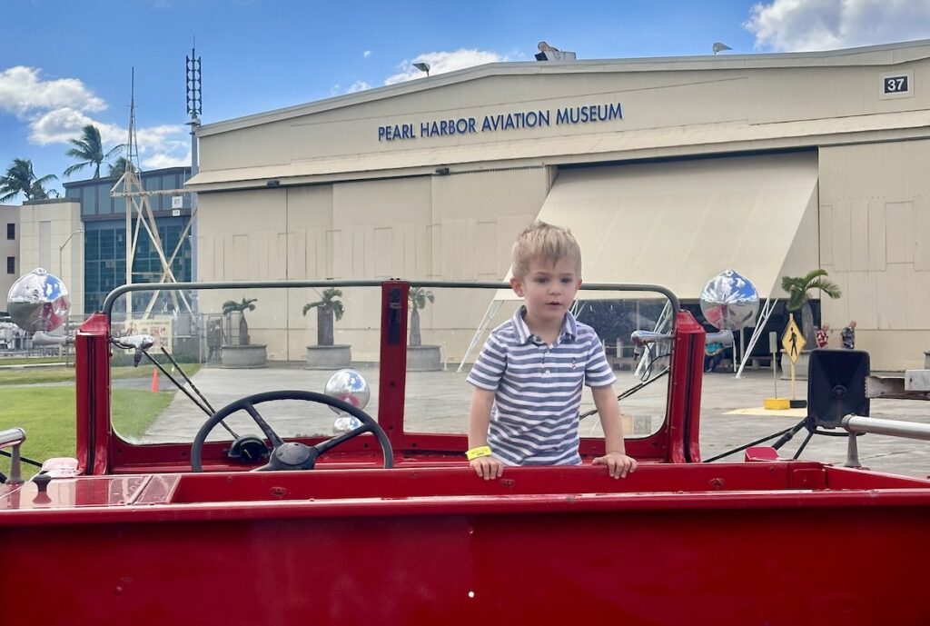 A young boy stands inside a bright red vintage fire truck, with the Pearl Harbor Aviation Museum visible in the background. The museum building is a beige structure with an open entrance. Pearl Harbor, Oahu.