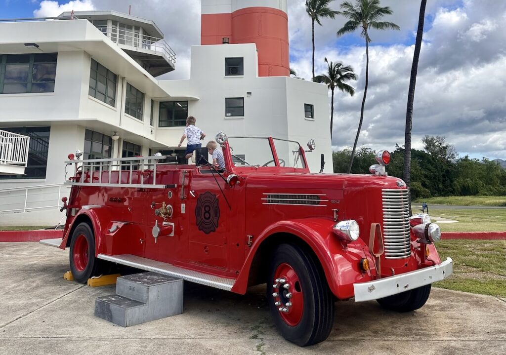 Two boys play in a bright red vintage fire truck. The truck is displayed in front of the Aviation Museum in Pearl Harbor, Oahu, Hawaii.