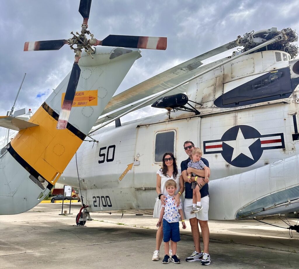 A family of four poses in front of a large military helicopter with visible propellers. The parents stand with their two young sons, who are smiling and enjoying the moment. Pearl Harbor Aviation Museum in Oahu.