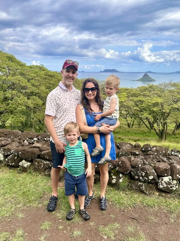 A family of four poses in front of a stone wall with Chinaman's Hat island in the distance, surrounded by greenery at Kualoa Ranch.