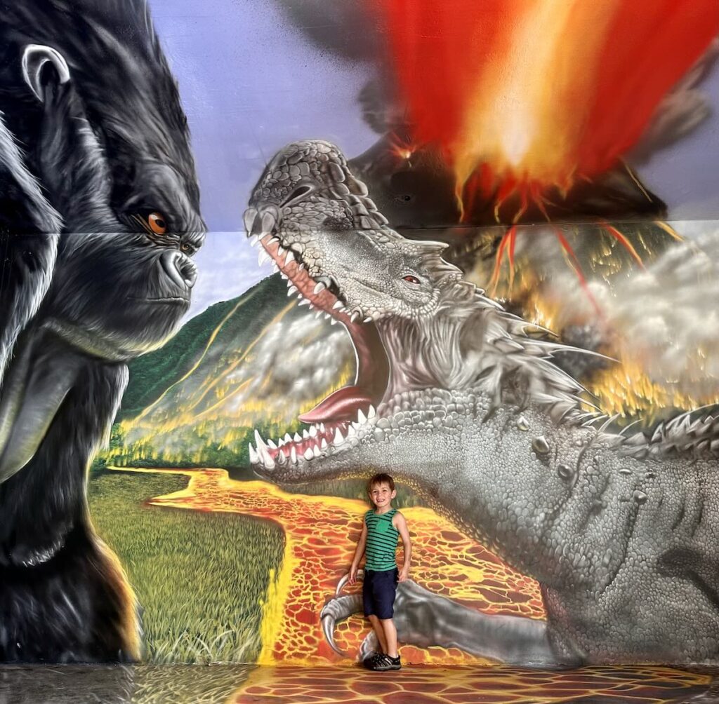 A young boy poses in front of a mural of a gorilla and a dinosaur roaring at each other, with lava and fire in the background. Kualoa Ranch, Oahu, Hawaii