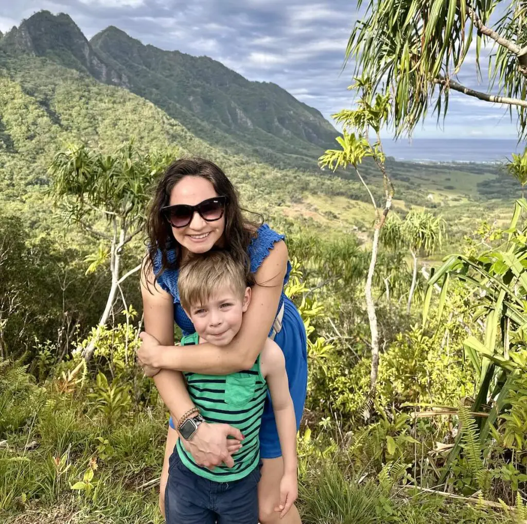 A mother and her son smile and hug in front of lush green mountains at Kualoa Ranch, surrounded by trees and plants under a partly cloudy sky. Oahu, Hawaii