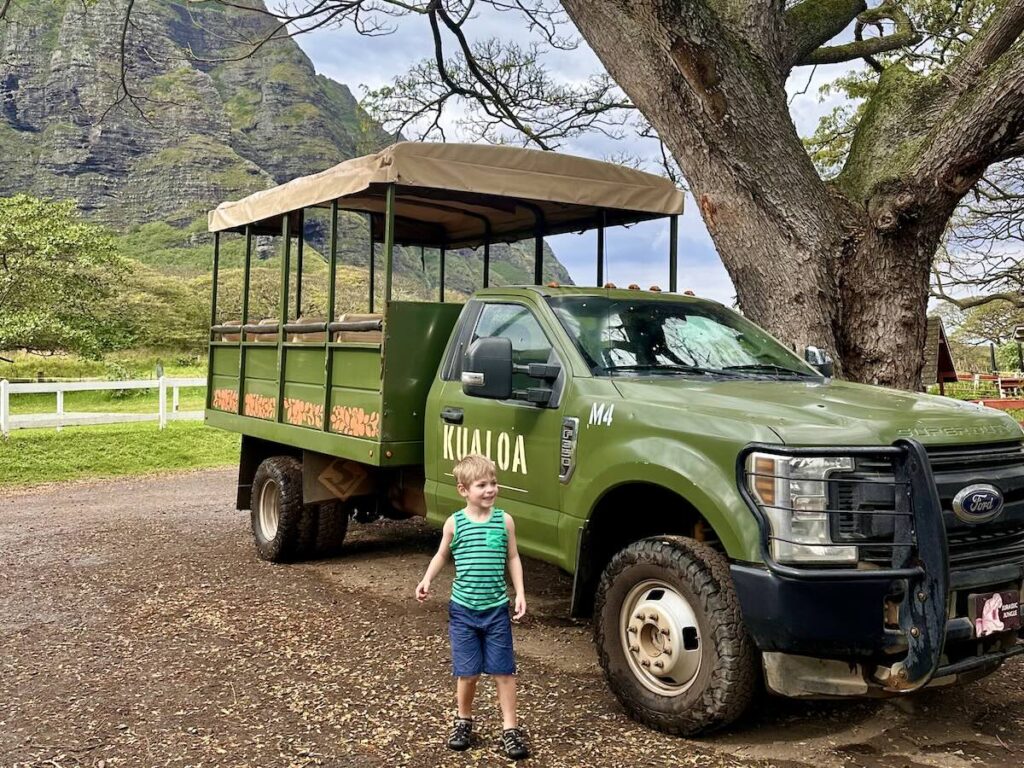 A young boy stands in front of a large green Kualoa Ranch truck, parked under a tree, with towering green mountains in the background.