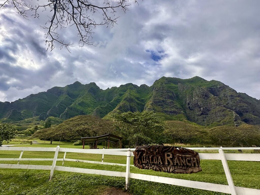 A white fence in front of lush green mountains at Kualoa Ranch, with a sign that says "Kualoa Ranch" and a cloudy sky above.