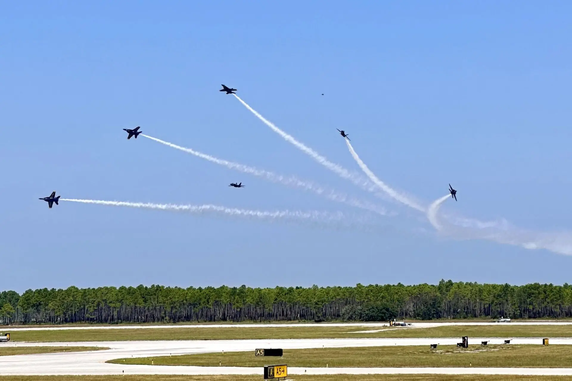 Six Blue Angels jets perform an aerial maneuver over the airfield, leaving white smoke trails in the sky as they spread apart. National Naval Aviation Museum, Pensacola, Florida