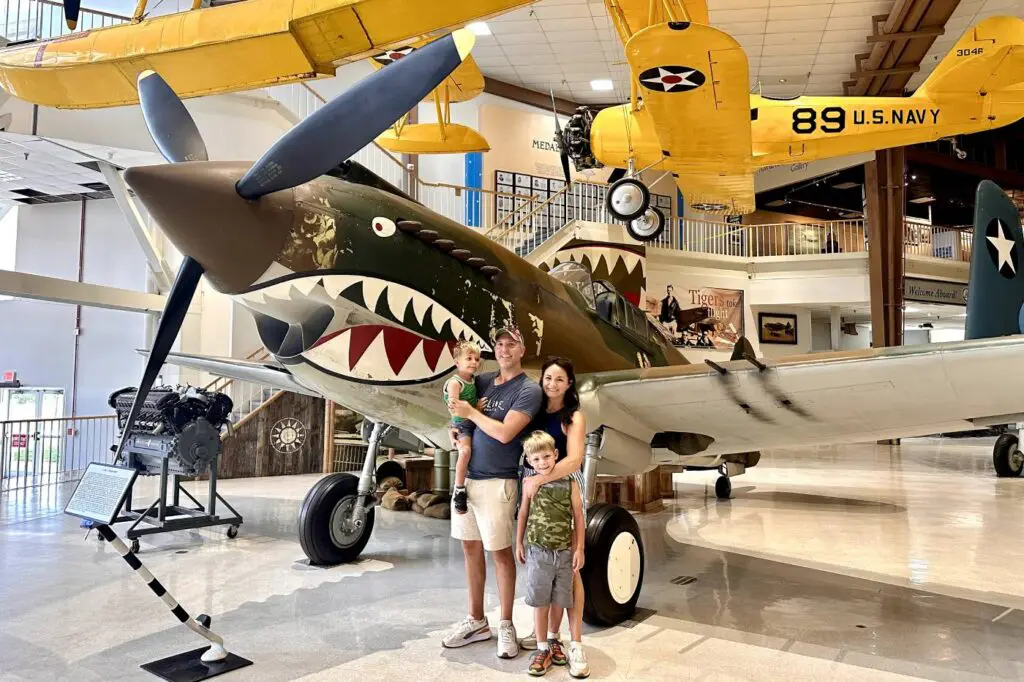 A family posing for a photo in front of a large vintage airplane exhibit inside the National Naval Aviation Museum in Pensacola, with the plane featuring a painted shark mouth.