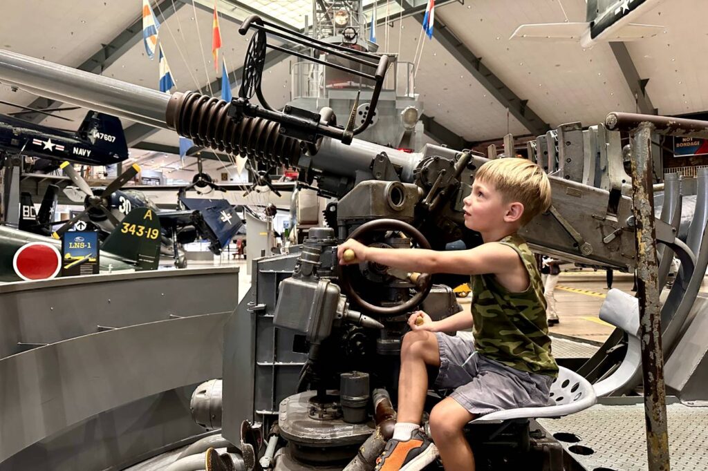 A young boy sits at the controls of a large, old naval artillery gun exhibit inside the National Naval Aviation Museum in Pensacola, concentrating as he moves the equipment.