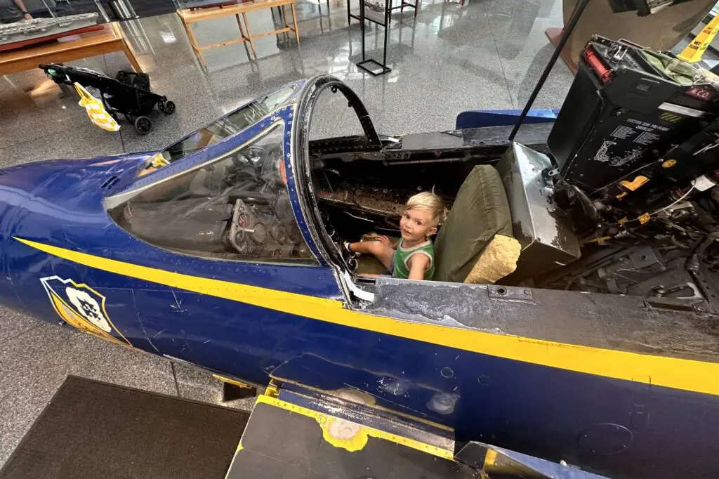 A young child sits inside the cockpit of a Blue Angels fighter jet on display, looking up while surrounded by the plane's complex controls. National Naval Aviation Museum, Pensacola, Florida