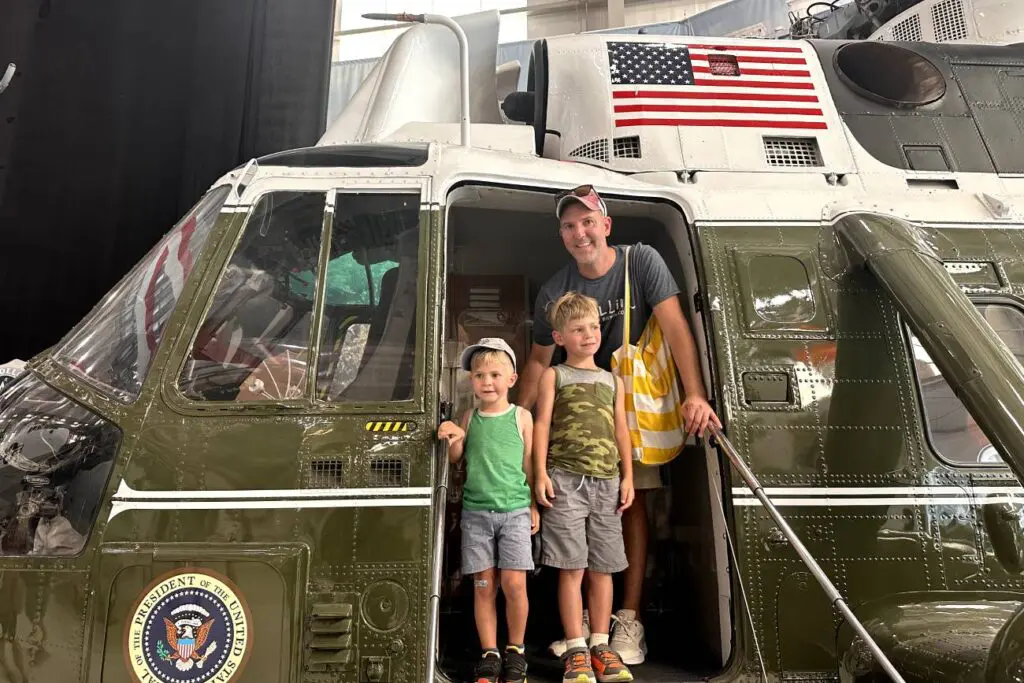 A father and two young boys stand inside a helicopter exhibit at the National Naval Aviation Museum in Pensacola, smiling and posing in front of the presidential seal and an American flag.