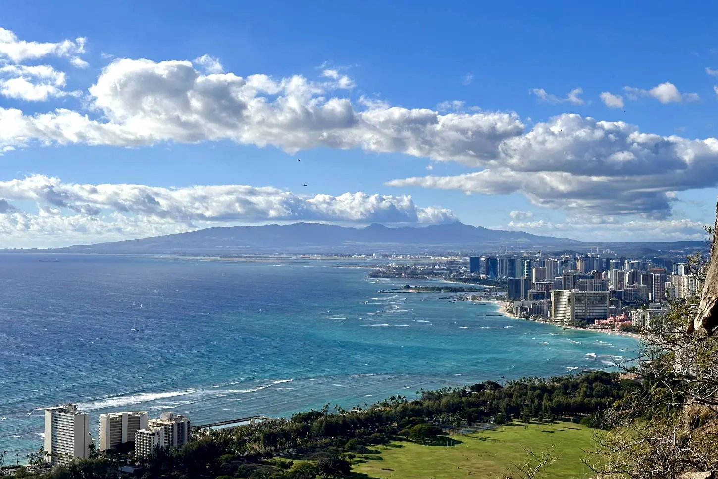 A breathtaking view from the Diamond Head summit in Oahu, showing the turquoise waters of the Pacific Ocean, the coastline of Waikiki with its high-rise buildings, and distant mountains under a bright blue sky with a few clouds.