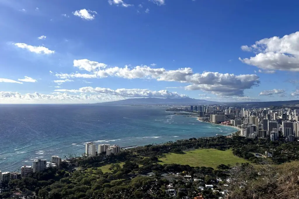 A view from the Diamond Head summit in Oahu, showing the turquoise waters of the Pacific Ocean, the coastline of Waikiki with its high-rise buildings, and distant mountains under a bright blue sky with a few clouds.