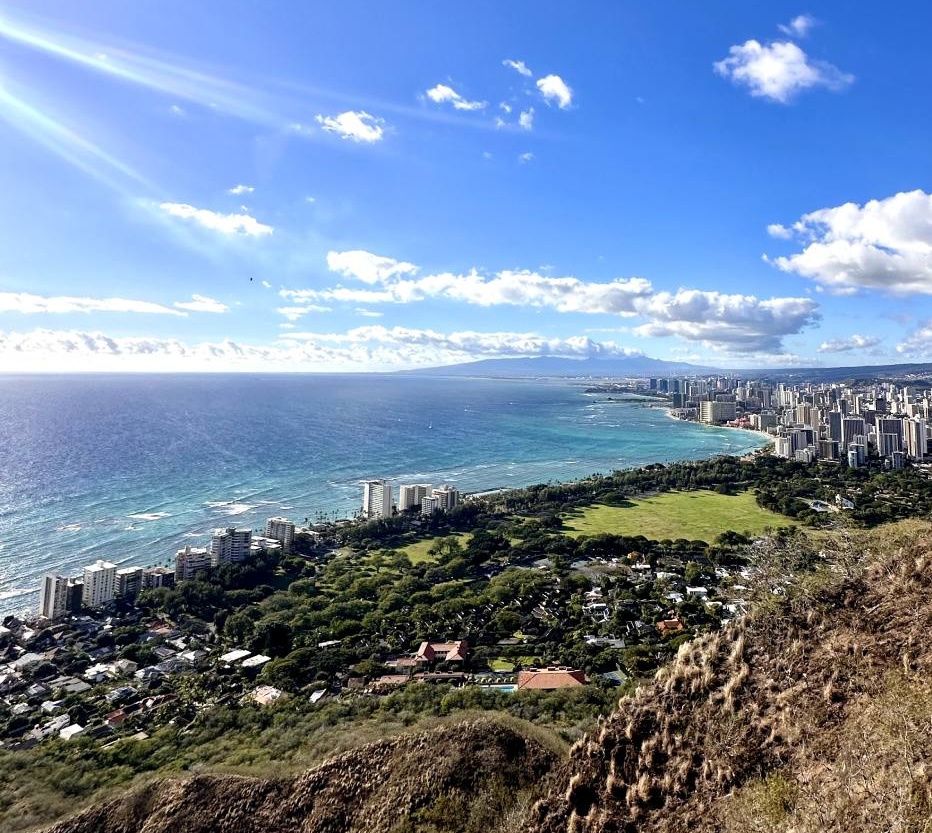 A wide-angle view from the Diamond Head summit in Oahu, capturing the lush green landscape below, the blue expanse of the Pacific Ocean, and the skyline of Waikiki under a clear, bright blue sky with a few clouds.