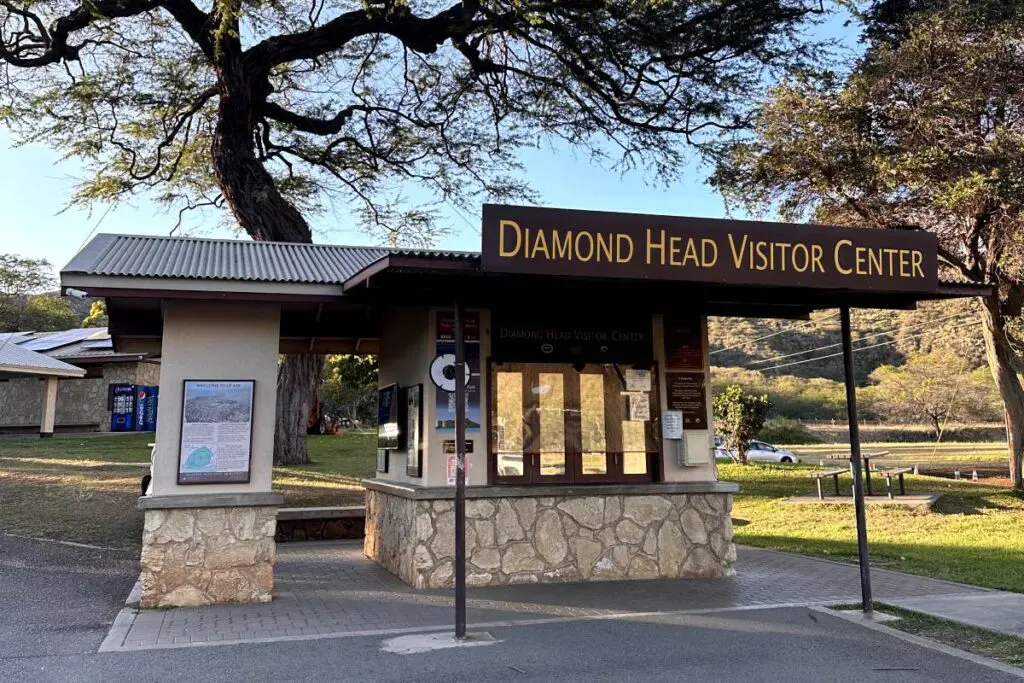 The Diamond Head Visitor Center in Oahu, a small building with a stone base and a sign above the entrance, stands in front of a large tree and grassy area. Nearby, vending machines are visible.