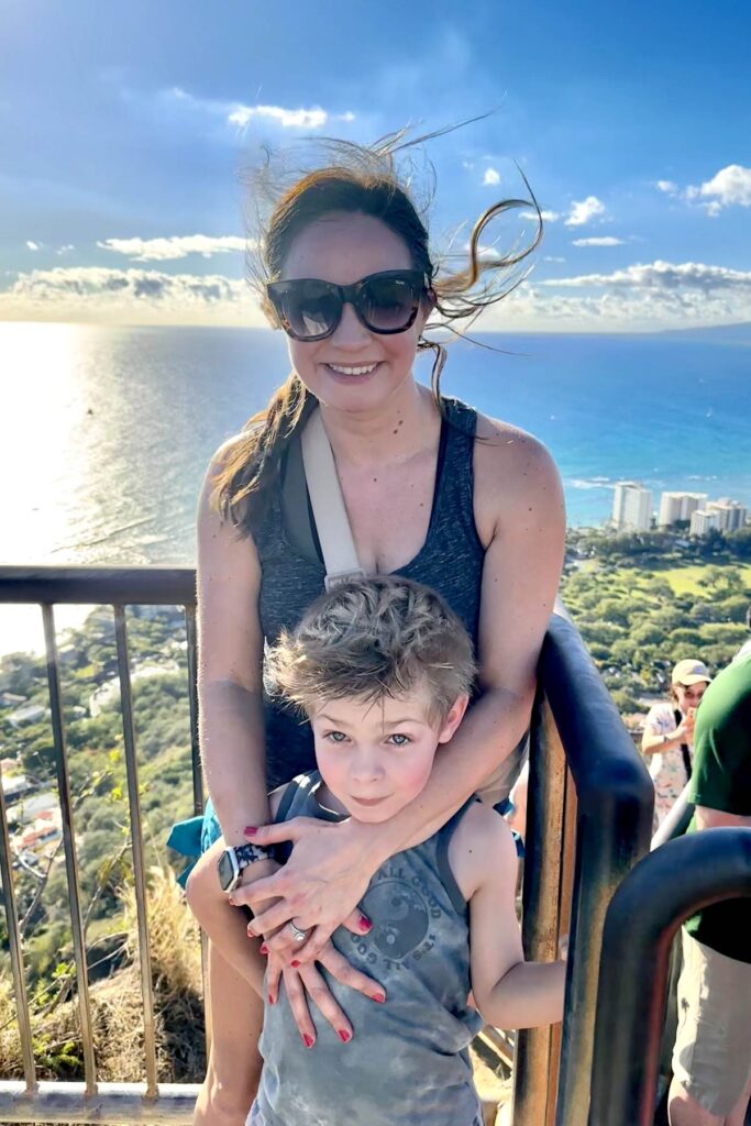 A smiling woman wearing sunglasses stands behind a young boy, hugging him from behind at the Diamond Head summit in Oahu. The ocean and Waikiki skyline are visible in the background under a bright blue sky.