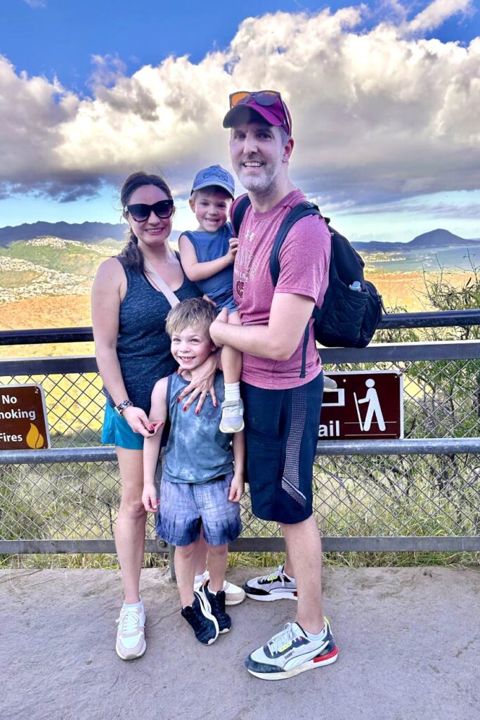 A family of four smiles for a photo at the Diamond Head summit in Oahu, standing in front of a fence with a scenic view of mountains and valleys in the background. The parents are holding their two young children, and everyone looks happy after completing the hike.