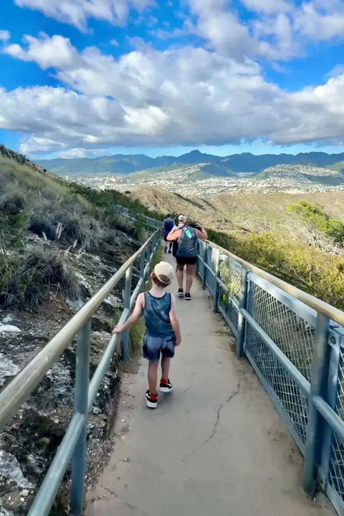 A child walks along a narrow, paved trail with metal guardrails, following behind an adult. The trail offers views of distant mountains and a valley under a blue sky with scattered clouds. Diamond Head, Oahu, Hawaii.