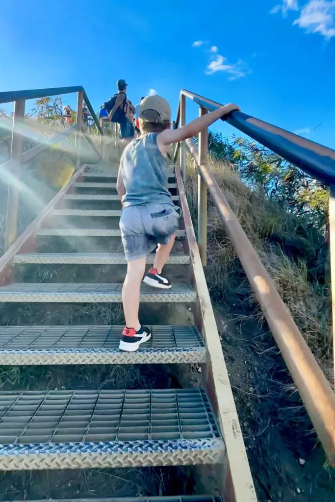 A child climbs a steep metal staircase, holding onto the handrail, with a bright blue sky and greenery visible in the background. Other hikers can be seen at the top of the stairs. Diamond Head, Oahu, Hawaii.