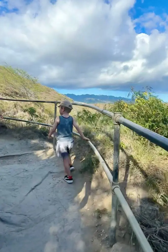 A child walks along a dirt trail with metal guardrails, holding onto the railing for balance. Diamond Head State Monument, Oahu, Hawaii.