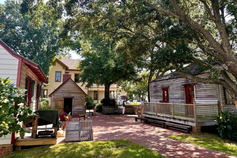 A peaceful courtyard in a historic village, surrounded by wooden buildings and large shady trees with brick walkways. Historic Pensacola Village, Florida