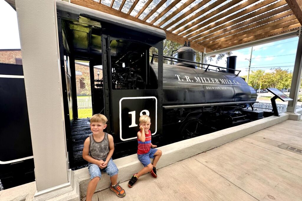 Two young boys sit in front of a black train engine labeled "T. R. Miller Mill Co." at an outdoor exhibit. Museum of Industry, Pensacola, Florida.